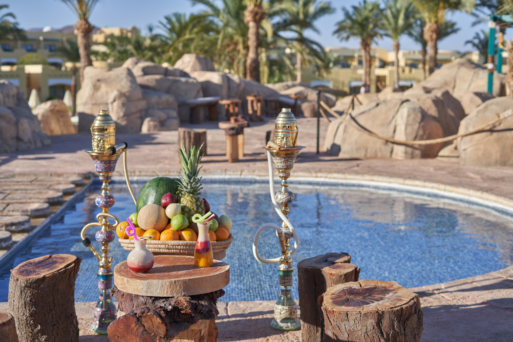 A big fruit platter, two sets of hookah, and fresh juice on a wooden table in front of a pool. In the background are sunny skies and palm trees.