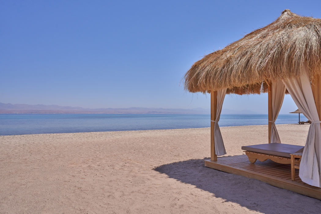 A view of shaded seating on a secluded beach with clear blue waters and mountains on the other side.