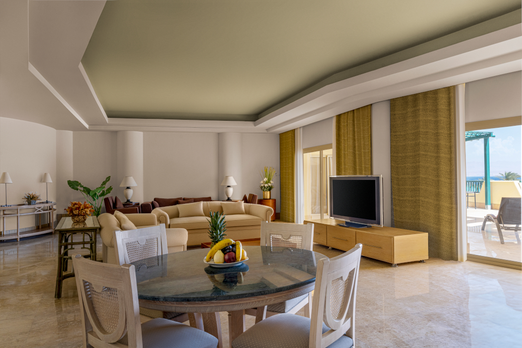 A sunny picture of the presidential suite living space showing a seating area around a flat-screen TV in the backrgound and a breakfast table with 4 chairs and a fruit platter in the foreground.