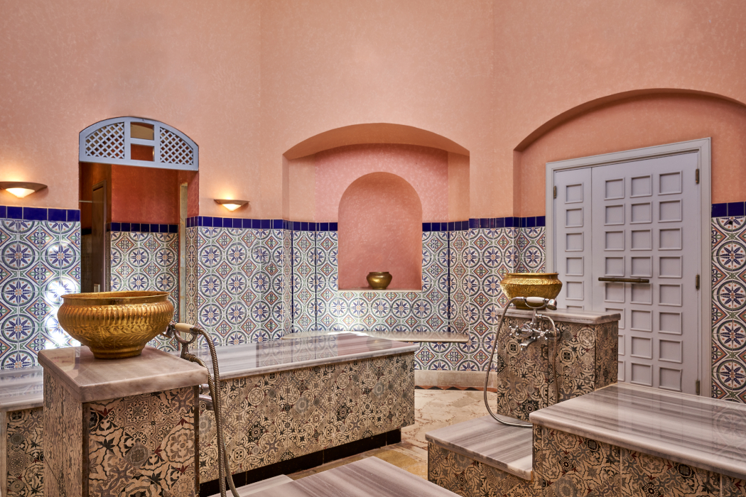 A picture showing a spa hammam setup with marble slabs and gold sinks. The walls are decorated in blue and orange patterns.